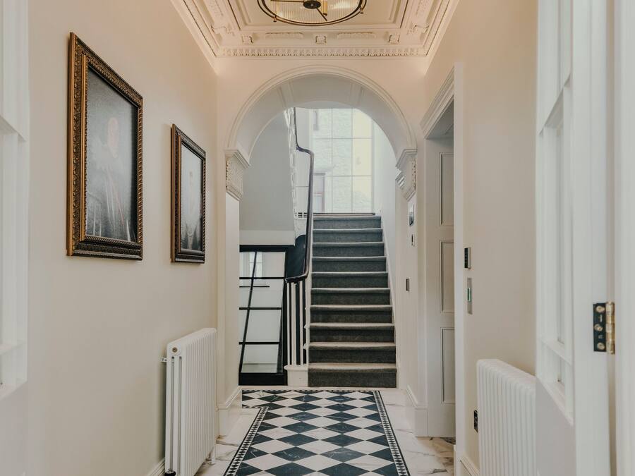 Un pasillo con suelo a cuadros blanco y negro, un candelabro, obras de arte enmarcadas en las paredes y una escalera que sube a otro piso.