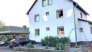 A two-story house with multiple windows, a covered porch, and a driveway with parked cars. - Cosy apartment for 2 guests with WIFI, TV and terrace (Stadtoldendorf)