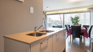 A modern kitchen with a double sink, a dining area with a table and chairs, and a large window with blinds. - 1 bedroom accommodation in Sint-Idesbald (Sint-Idesbald)