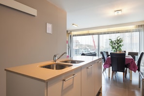 Een moderne keuken met een dubbele spoelbak, een zithoek met een tafel en stoelen, en een groot raam met zonwering.