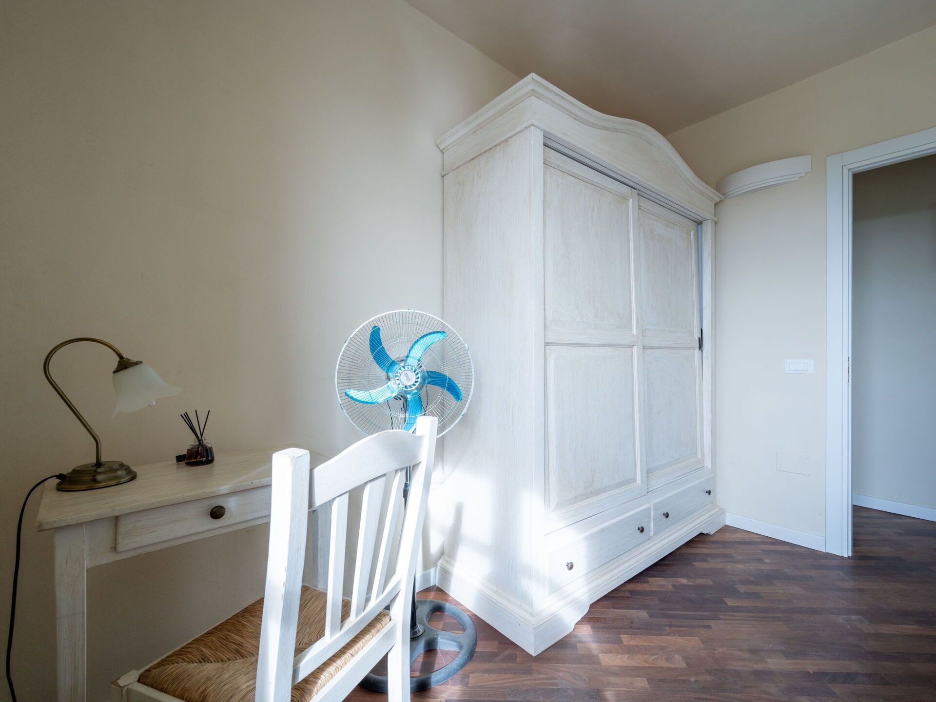 Ein Zimmer mit einem weißen Kleiderschrank, einem Schreibtisch mit Lampe und einem Standventilator.