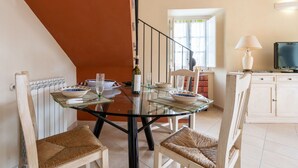 Un coin repas avec une table en verre, des chaises en osier et une vue sur une cuisine équipée d’un téléviseur et de livres.