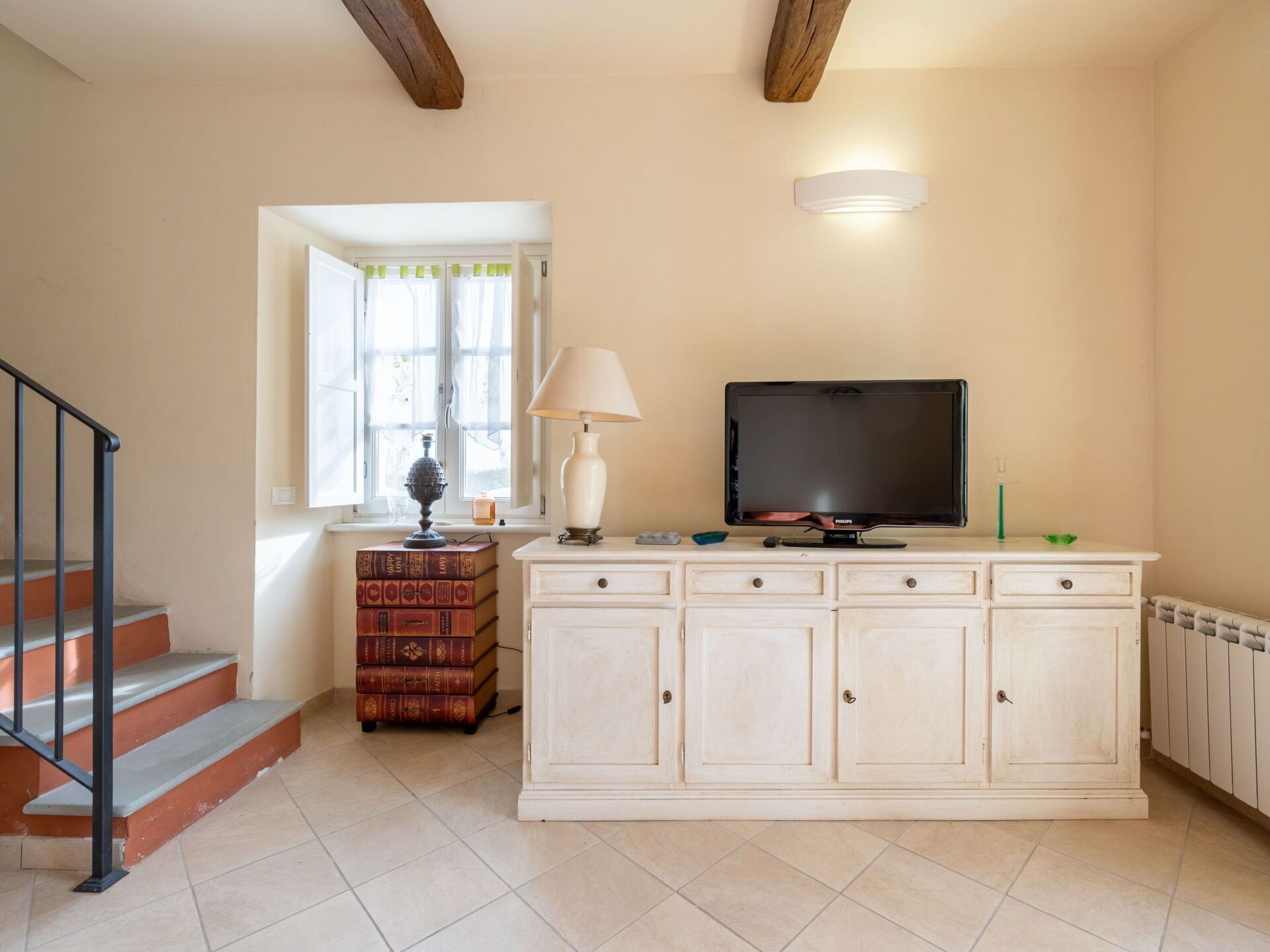 Un salon avec un escalier, un téléviseur, un meuble et une fenêtre avec des rideaux.