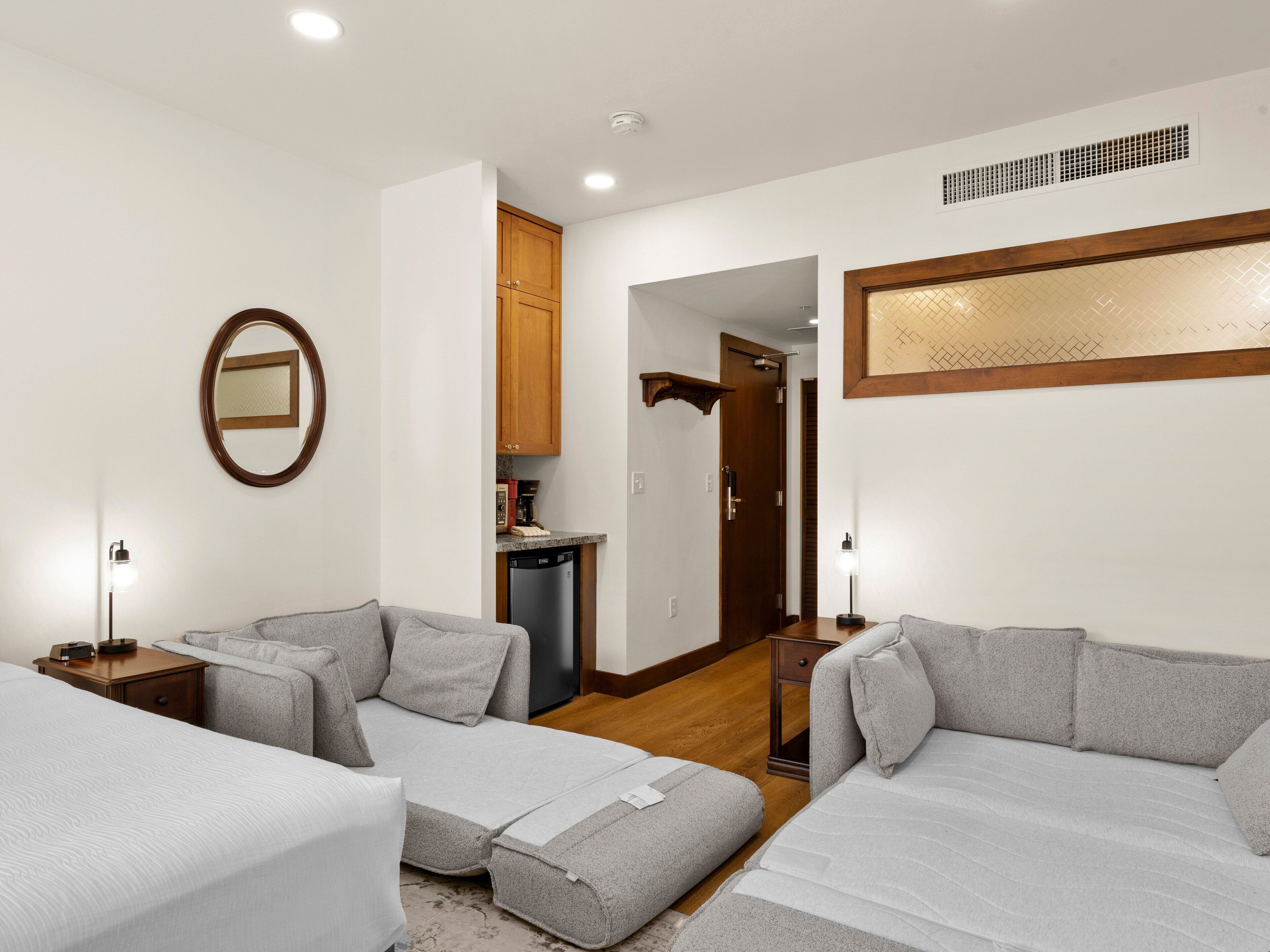 Habitación de hotel con dos camas, una kitchenette pequeña, un espejo y una puerta que da a otra habitación.