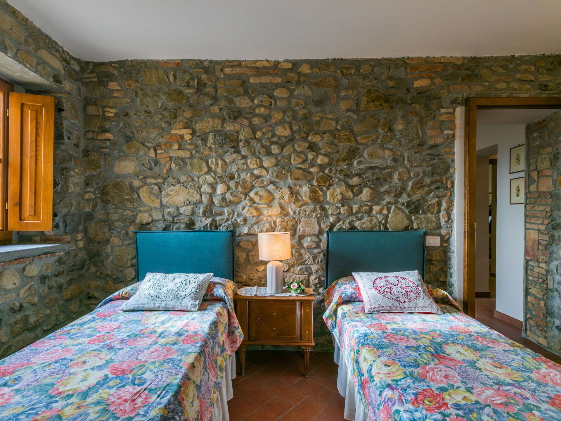 Une chambre avec deux lits, chacun agrémenté de draps à motifs floraux, un mur en pierre et une armoire en bois avec une lampe.