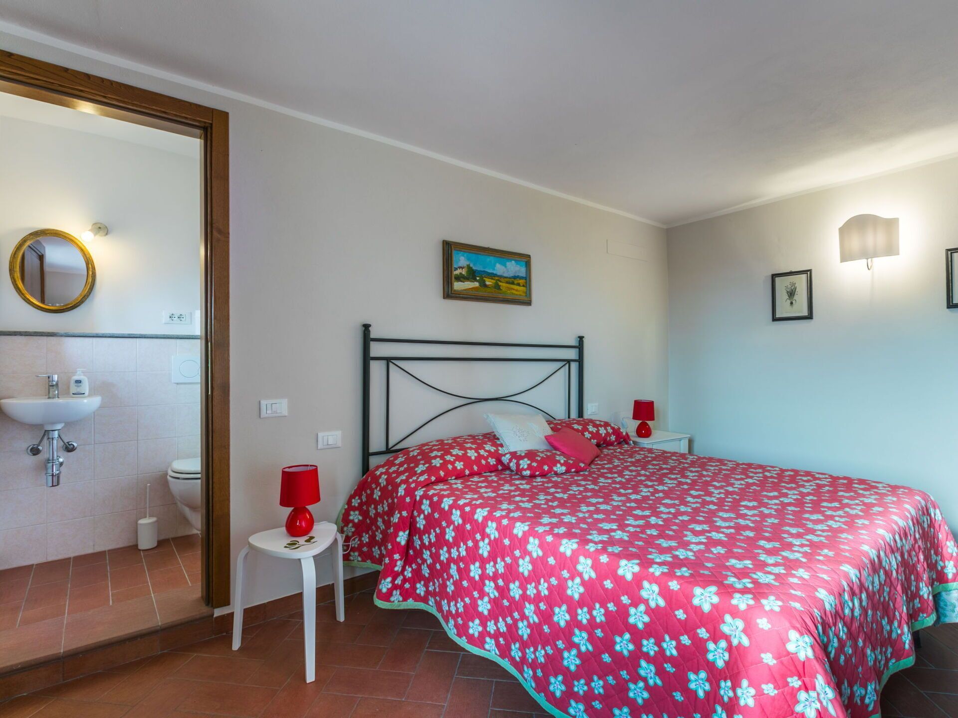 Un dormitorio con cama, mesita de noche, lámpara roja, un cuadro, y un baño con lavabo e inodoro.