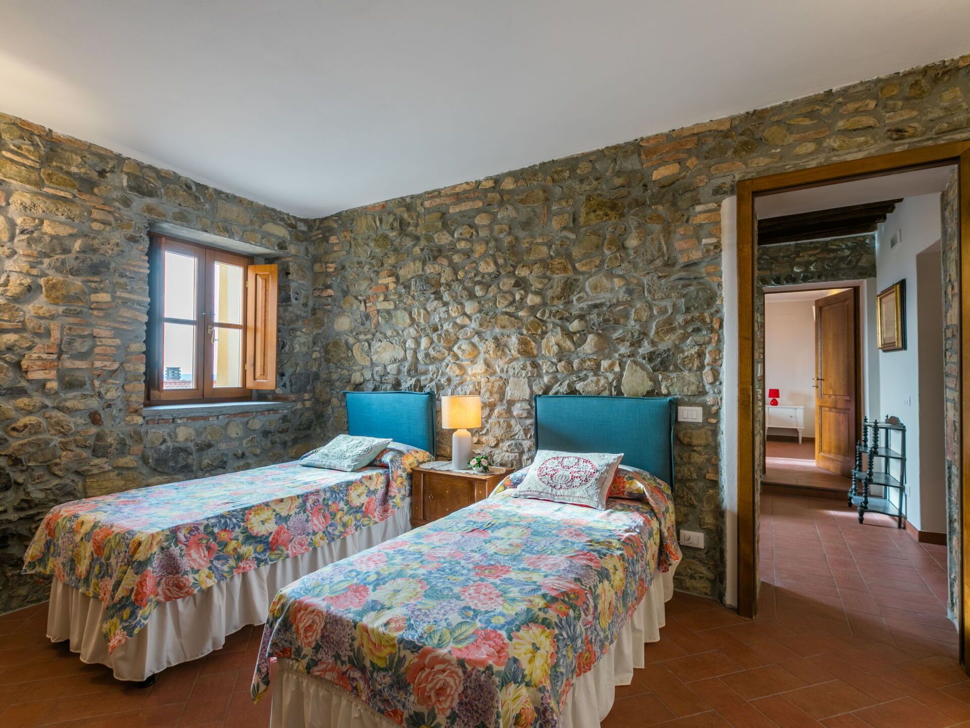 Una habitación con dos camas, cada una con ropa de cama floral, una pared de piedra, una puerta de madera y una ventana con contraventanas.