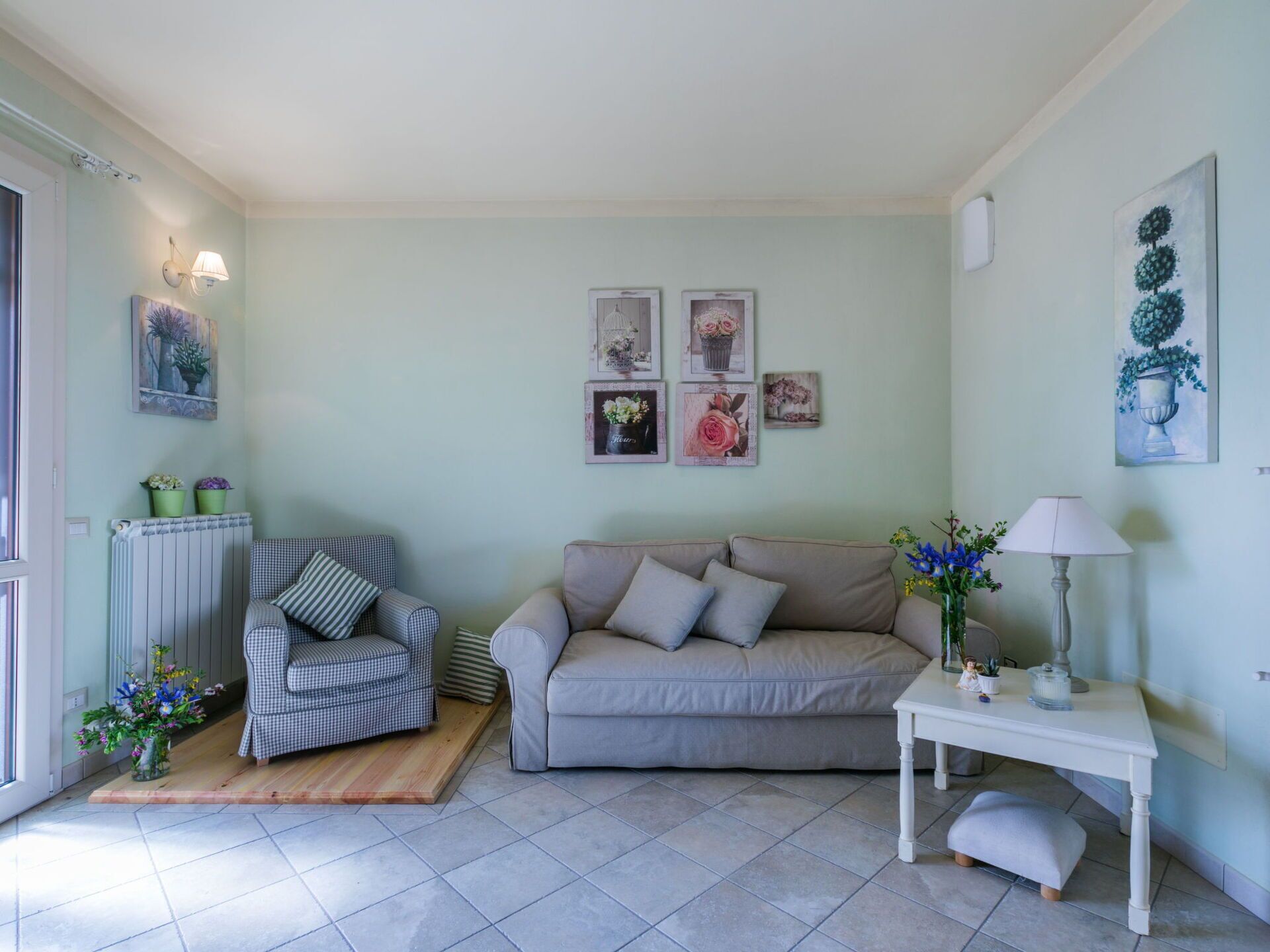 Una sala de estar con un sillón a cuadros, un sofá beige y una mesita auxiliar blanca con una lámpara. Se observan cuadros en la pared y un radiador a la izquierda.