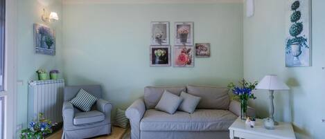 Una sala de estar con un sillón a cuadros, un sofá beige y una mesita auxiliar blanca con una lámpara. Se observan cuadros en la pared y un radiador a la izquierda.