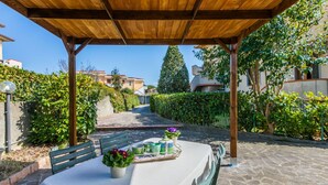 A covered outdoor dining area with a table set for two, surrounded by stone flooring and greenery.