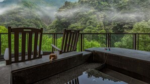 A balcony with a view of a lush forest and mist-covered mountains. There are two wooden chairs and a table with a wooden bowl and a cup on it. - Sunrise Hot Spring Hotel (Taian)