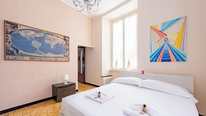 Habitación de hotel con un gran mapa del mundo, cama con ropa blanca, mesita de noche pequeña con lámpara y ventana con cortinas.