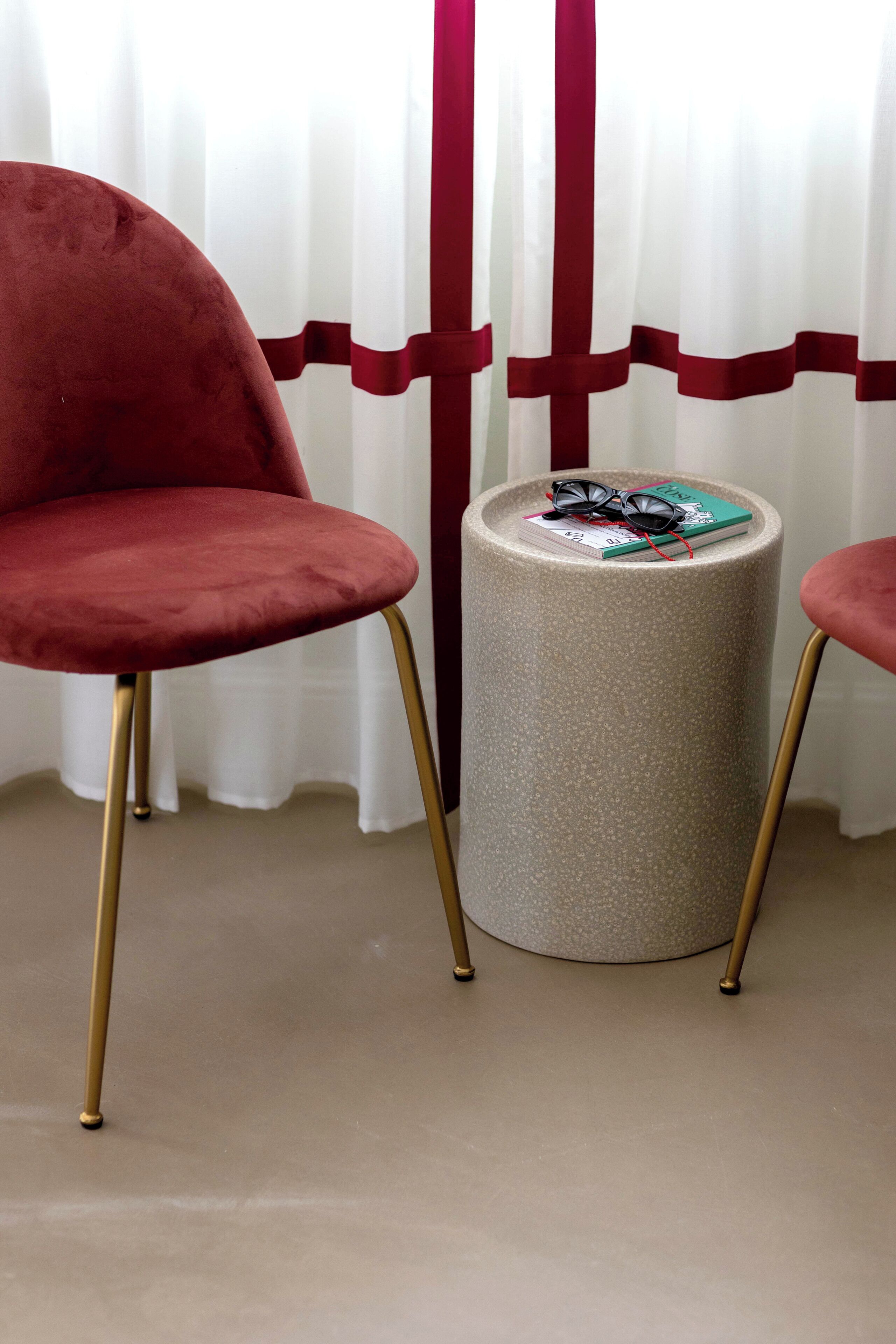 A red chair, a round table with a book and sunglasses on it, and a curtain with a red stripe.