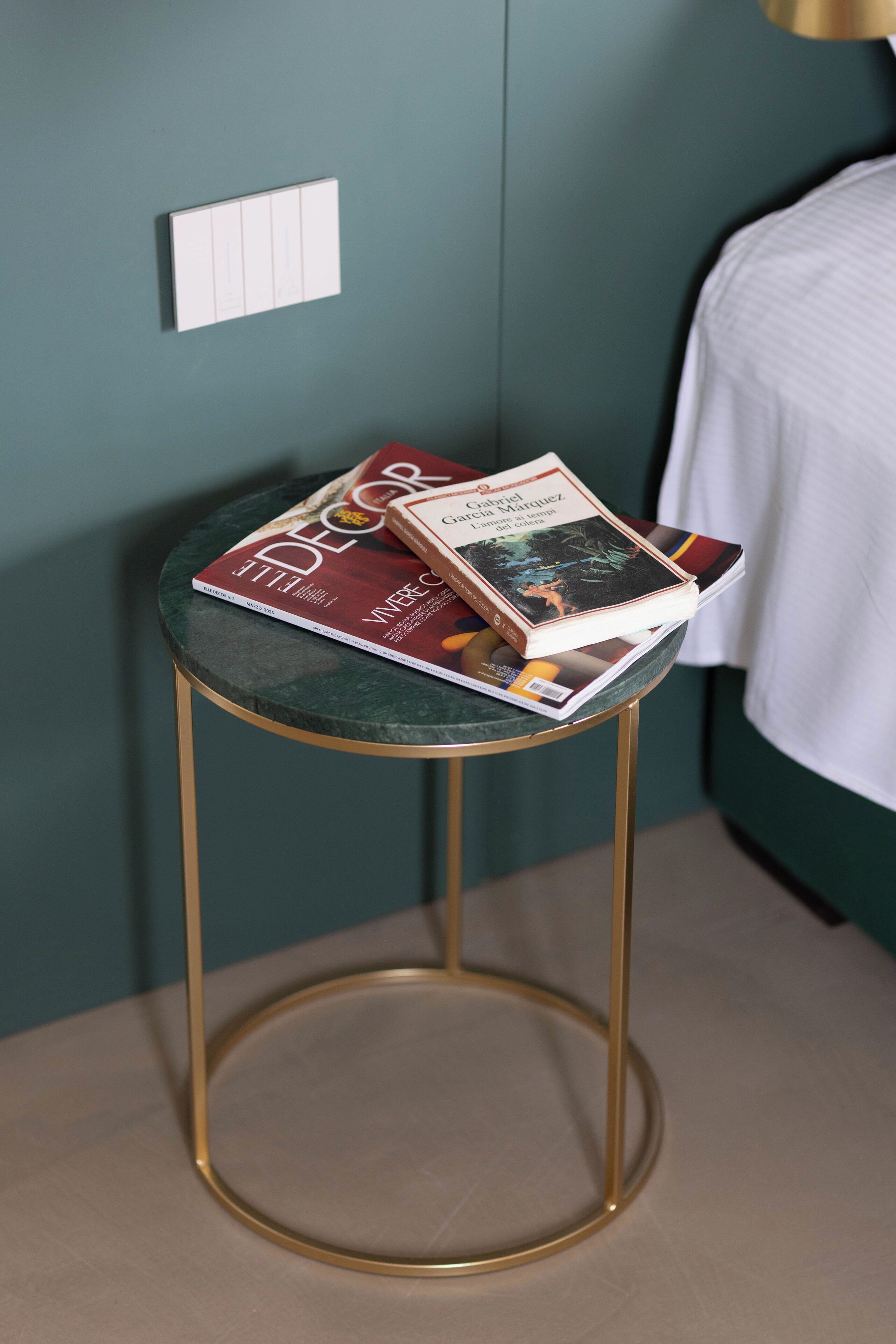 Un tavolino piccolo e rotondo con un piano in marmo verde e gambe dorate, su cui sono posizionati una rivista e un libro.