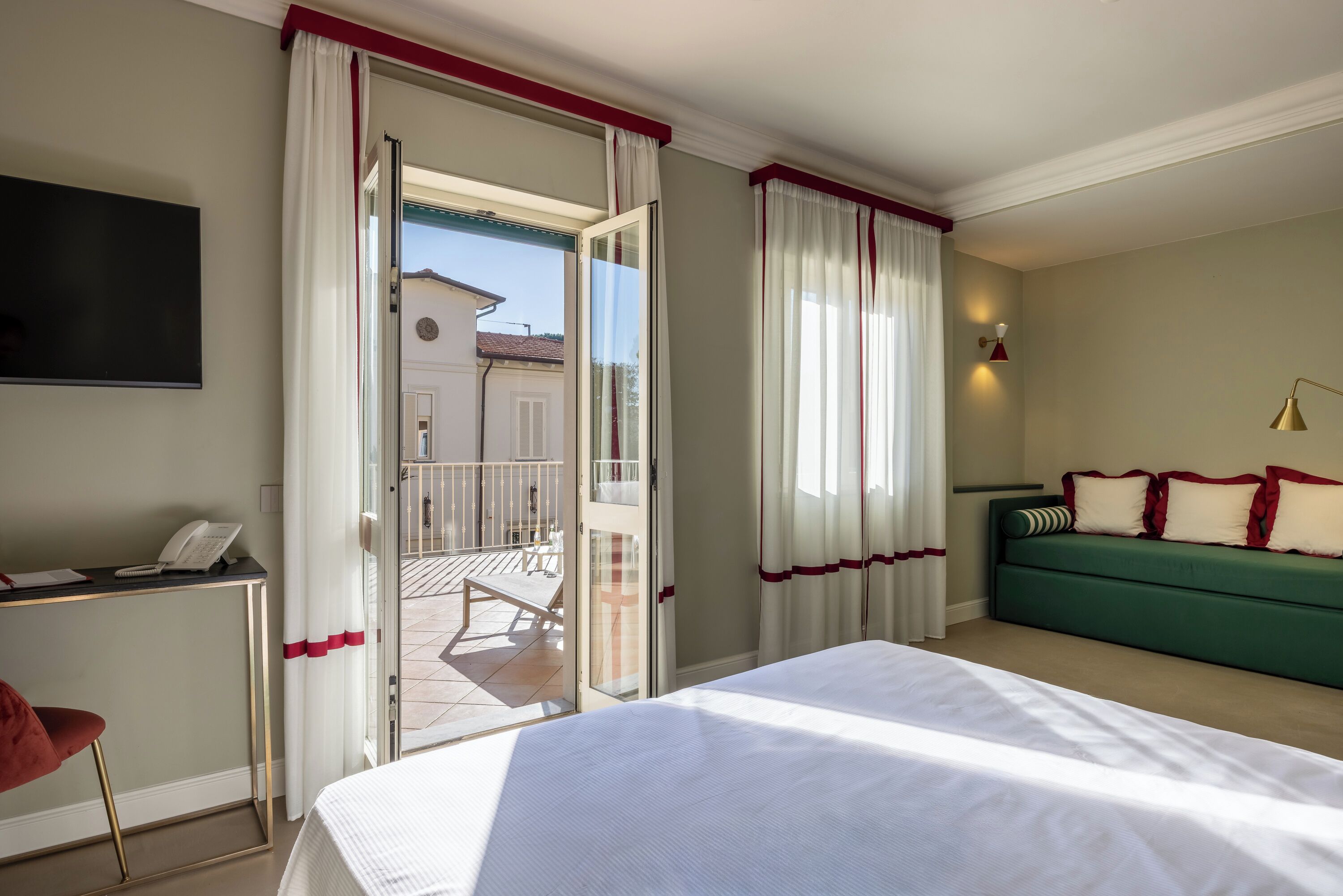 Ein Hotelzimmer mit Bett, Schreibtisch mit Telefon, grünem Sofa mit roten Kissen und Balkon mit Stuhl.