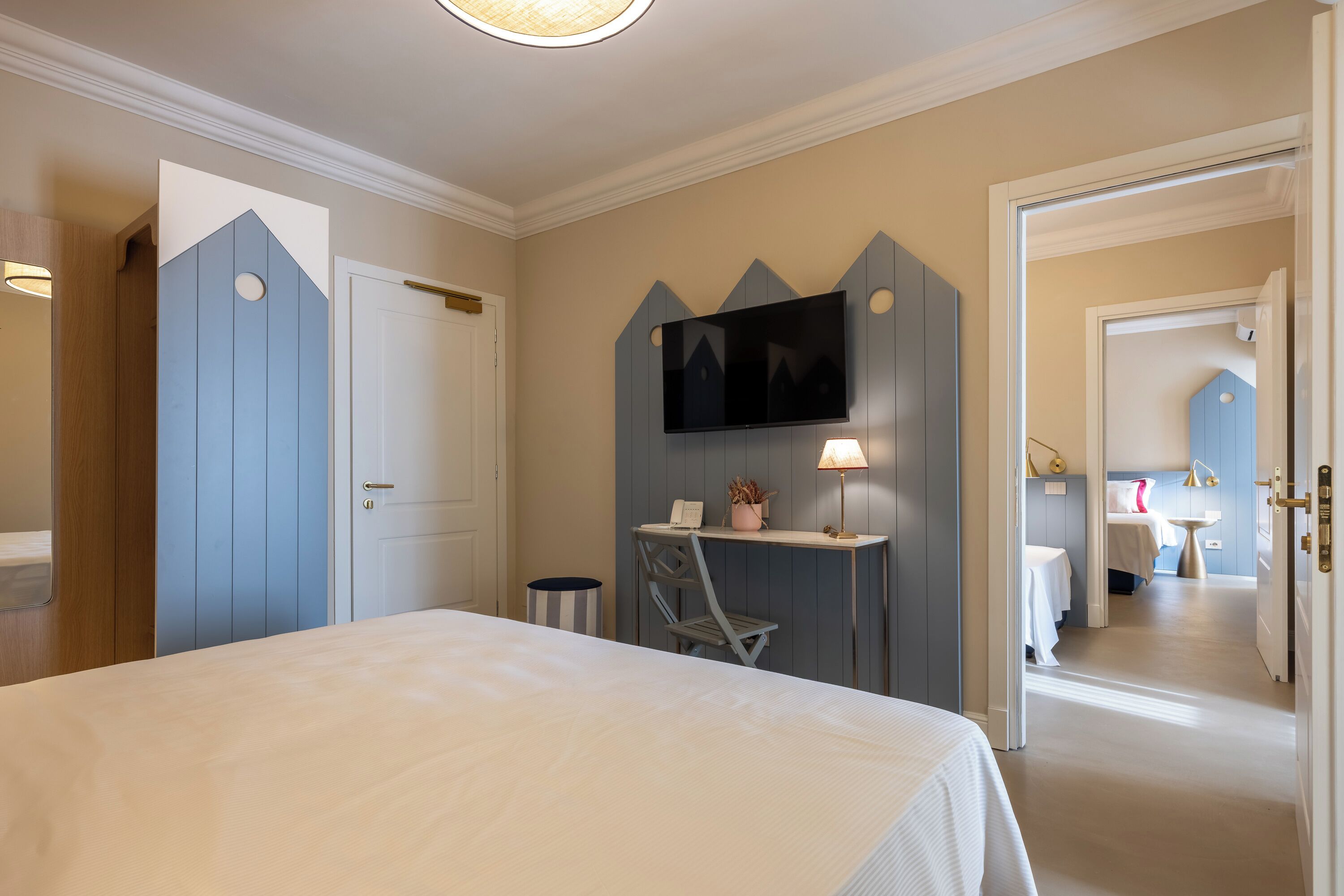A bedroom with a bed, a desk with a chair, a TV mounted on a wall, and a bathroom visible through an open door.