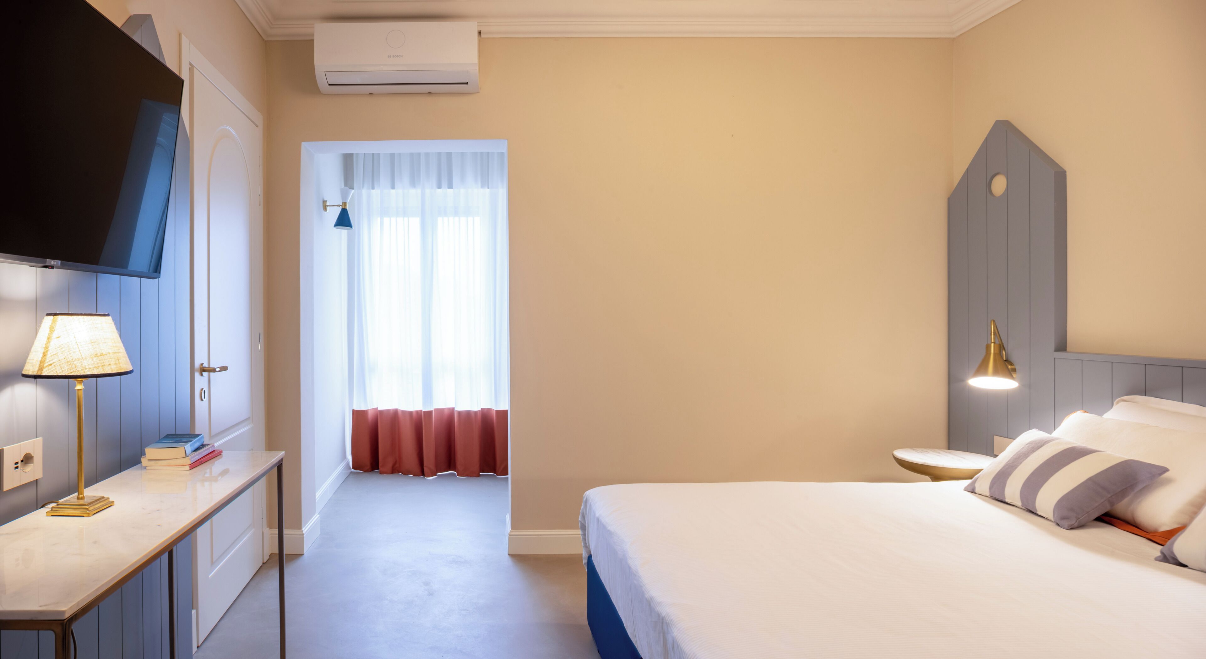 A hotel room with a bed, bedside table, lamp, TV, and a window with curtains.