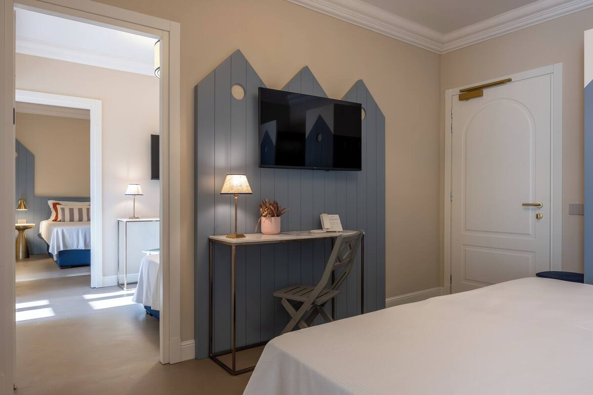 A hotel room with a bed, a desk with a lamp, a chair, a TV mounted on a wall, and a door.