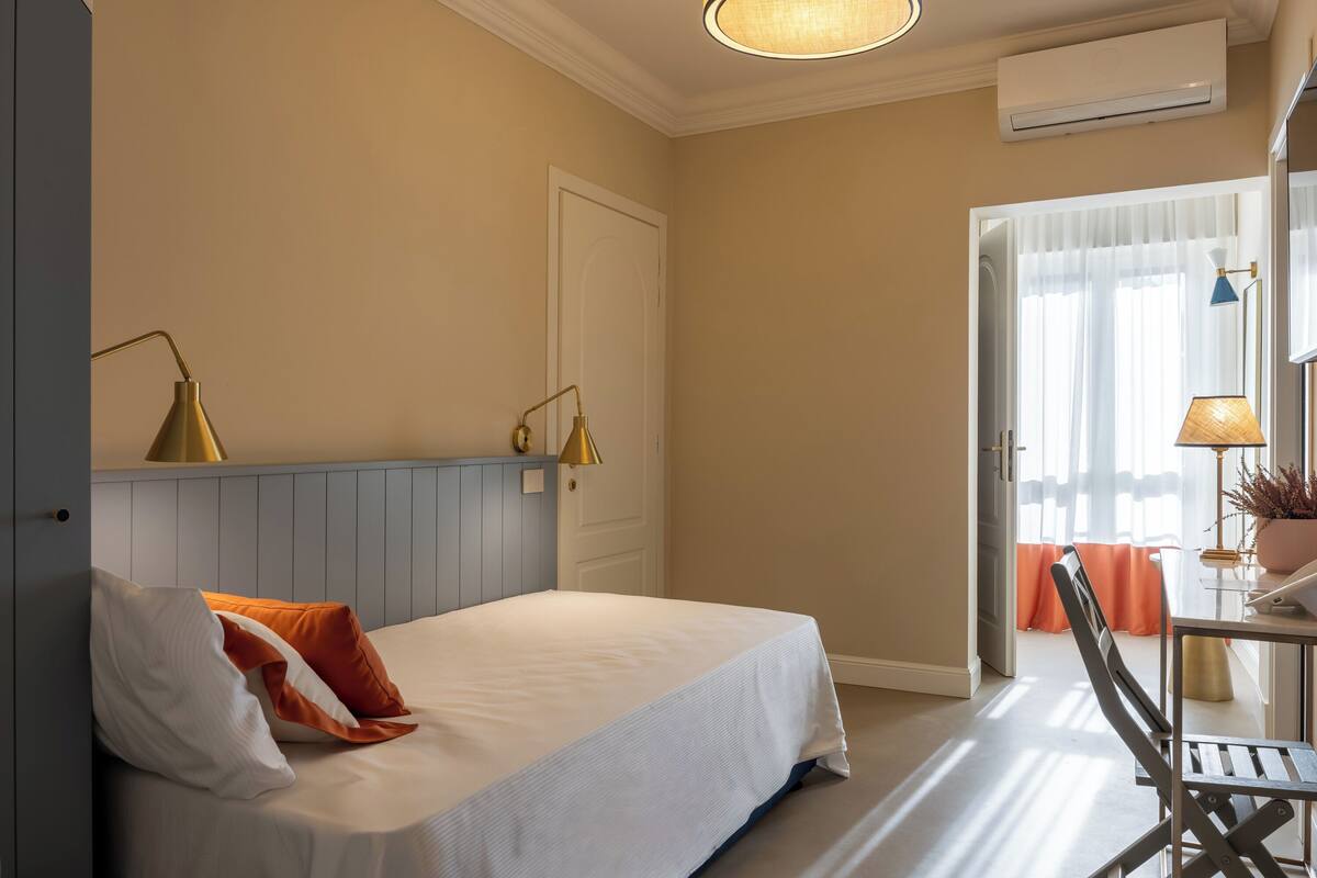 A hotel room with a bed, two bedside lamps, a desk with a chair, and a window with curtains.