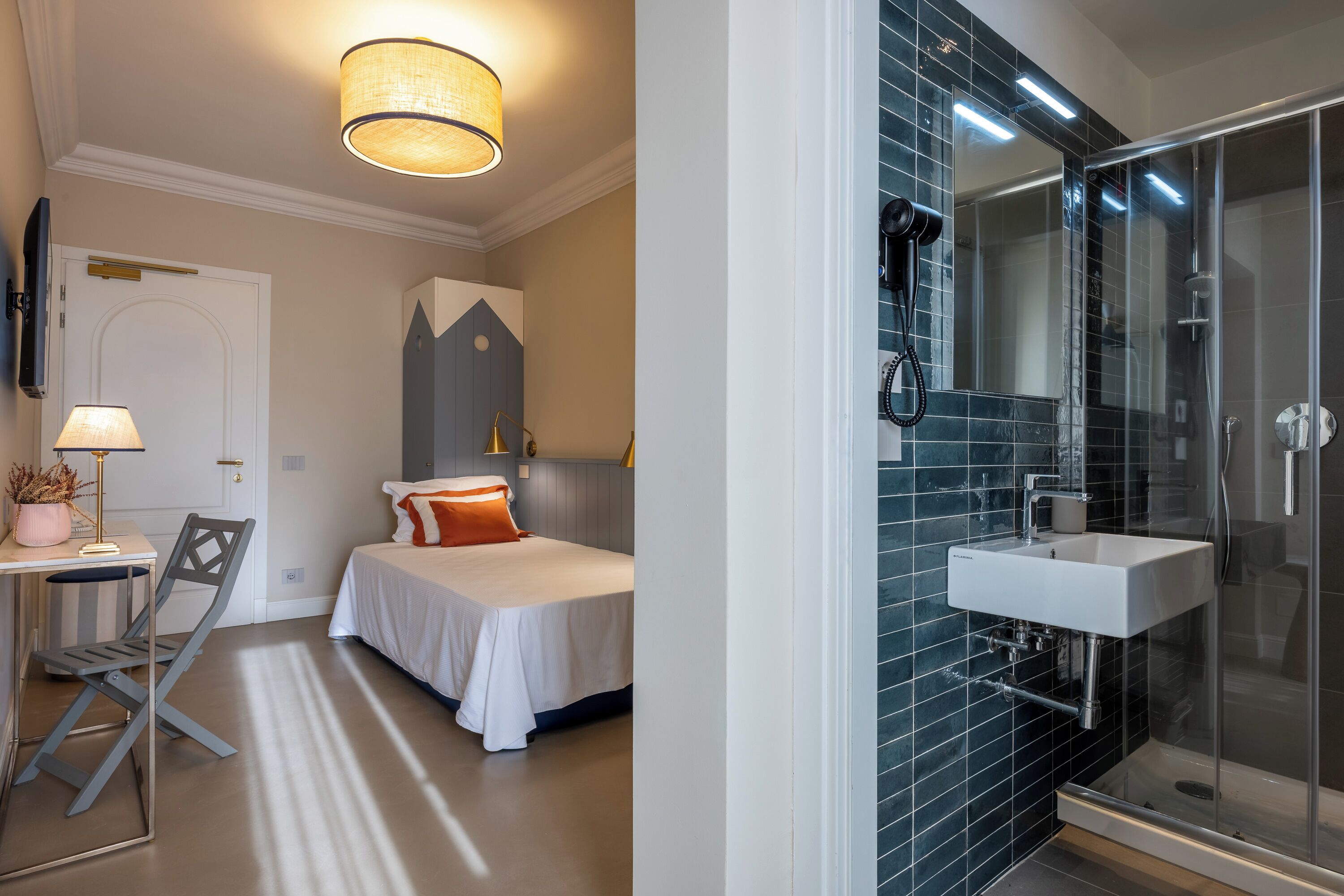 A bedroom with a bed, bedside table, lamp, and a chair. The bathroom features a glass shower enclosure, sink, and mirror.