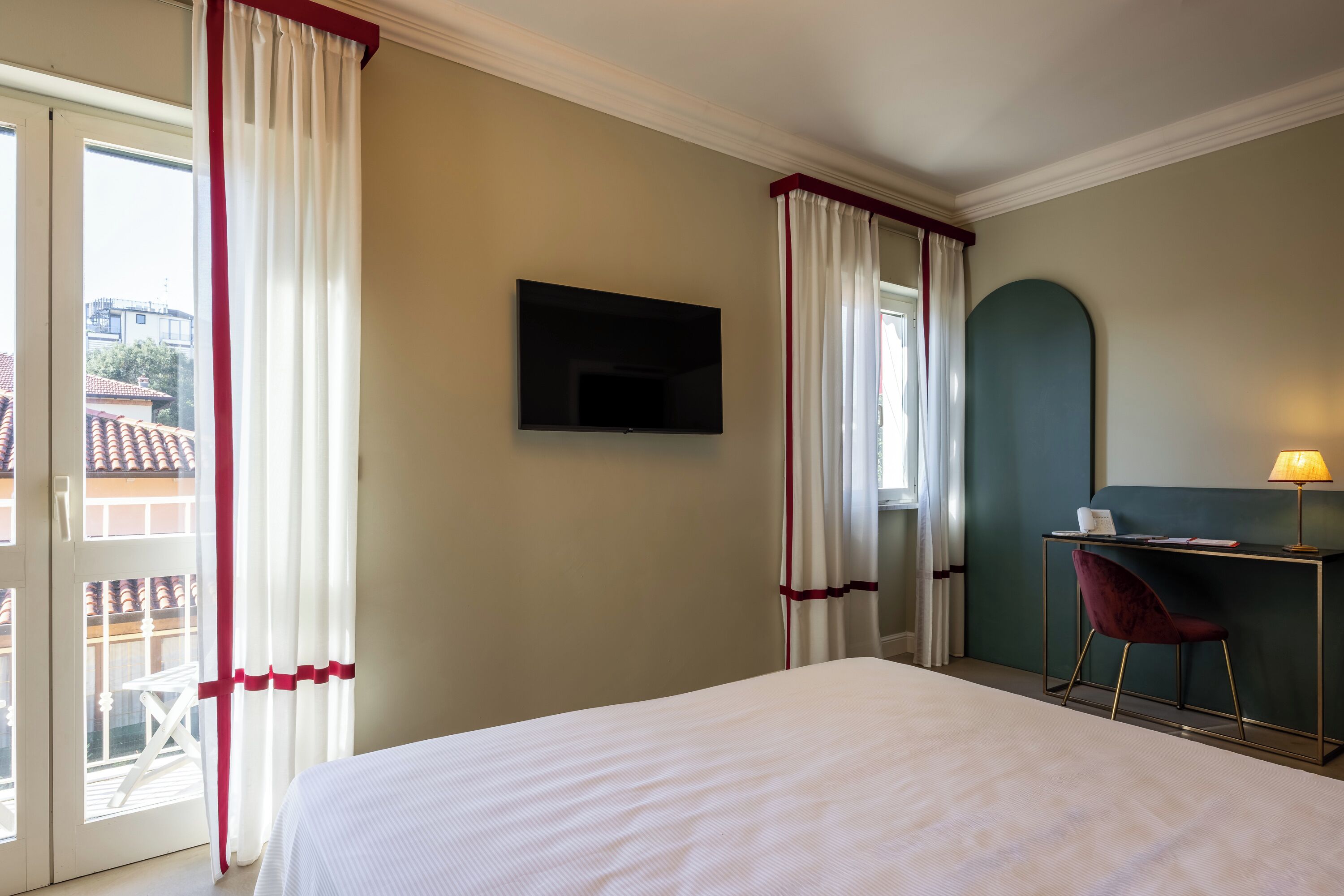 A hotel room with a bed, a desk with a chair, a TV, and a balcony with curtains.