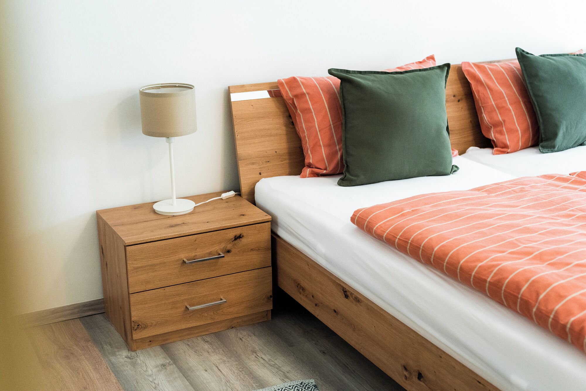 Ein ordentlich bezogenes Bett mit einer gestreiften Decke, ein Nachttisch aus Holz mit einer Lampe und zwei Schubladen.