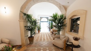 A spacious hallway with a checkered floor, two large potted plants, a beige armchair, a small round table, and a decorative vase.