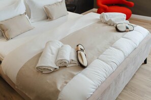 Una cama bien hecha con sábanas blancas, un par de zapatillas y una toalla doblada sobre un edredón de color claro.