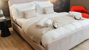 A neatly made bed with white linens and towels, a small round table with a bottle, and a red chair in a modern room with a stone wall and wooden floor.