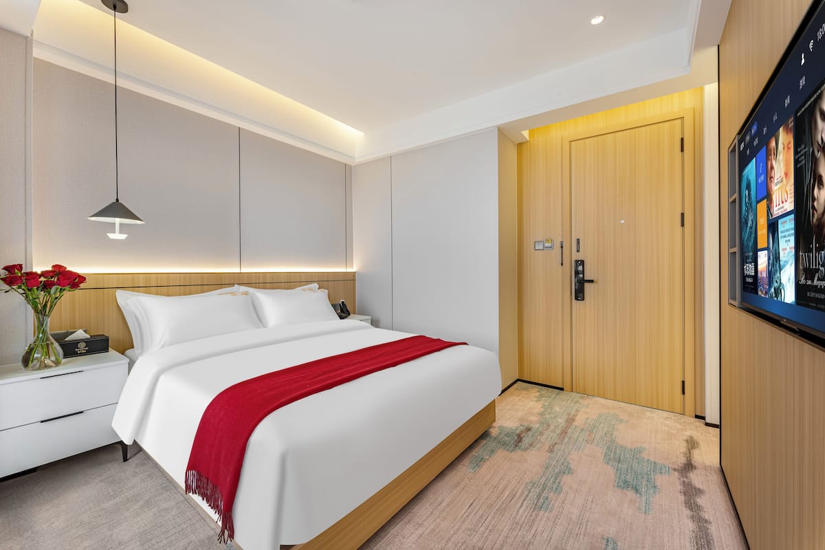 Een moderne hotelkamer met een groot bed, een nachtkastje met een vaas rode bloemen, een houten deur en een aan de muur bevestigde televisie.