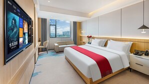 Een moderne hotelkamer met een grote flatscreen-tv waarop filmposters staan, een opgemaakt bed met een rode plaid, een nachtkastje met een telefoon en een bloemenvaas, een klein bureau met een stoel, en een groot raam met uitzicht over de stad.