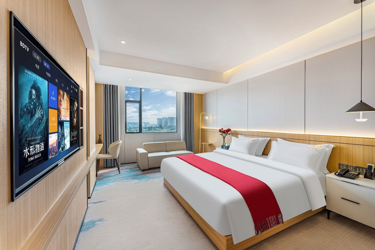 Een moderne hotelkamer met een grote flatscreen-tv waarop filmposters staan, een opgemaakt bed met een rode plaid, een nachtkastje met een telefoon en een bloemenvaas, een klein bureau met een stoel, en een groot raam met uitzicht over de stad.
