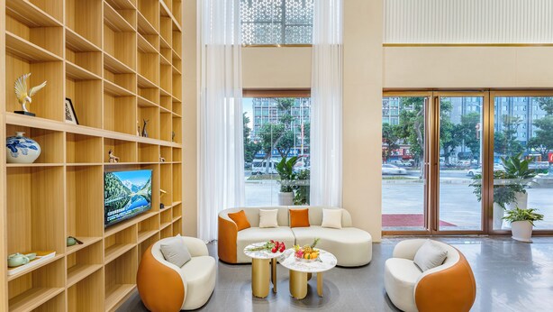 Een moderne woonkamer met een bank, twee fauteuils en een salontafel. Een televisie staat op een plank, en een groot raam biedt uitzicht op de stad.