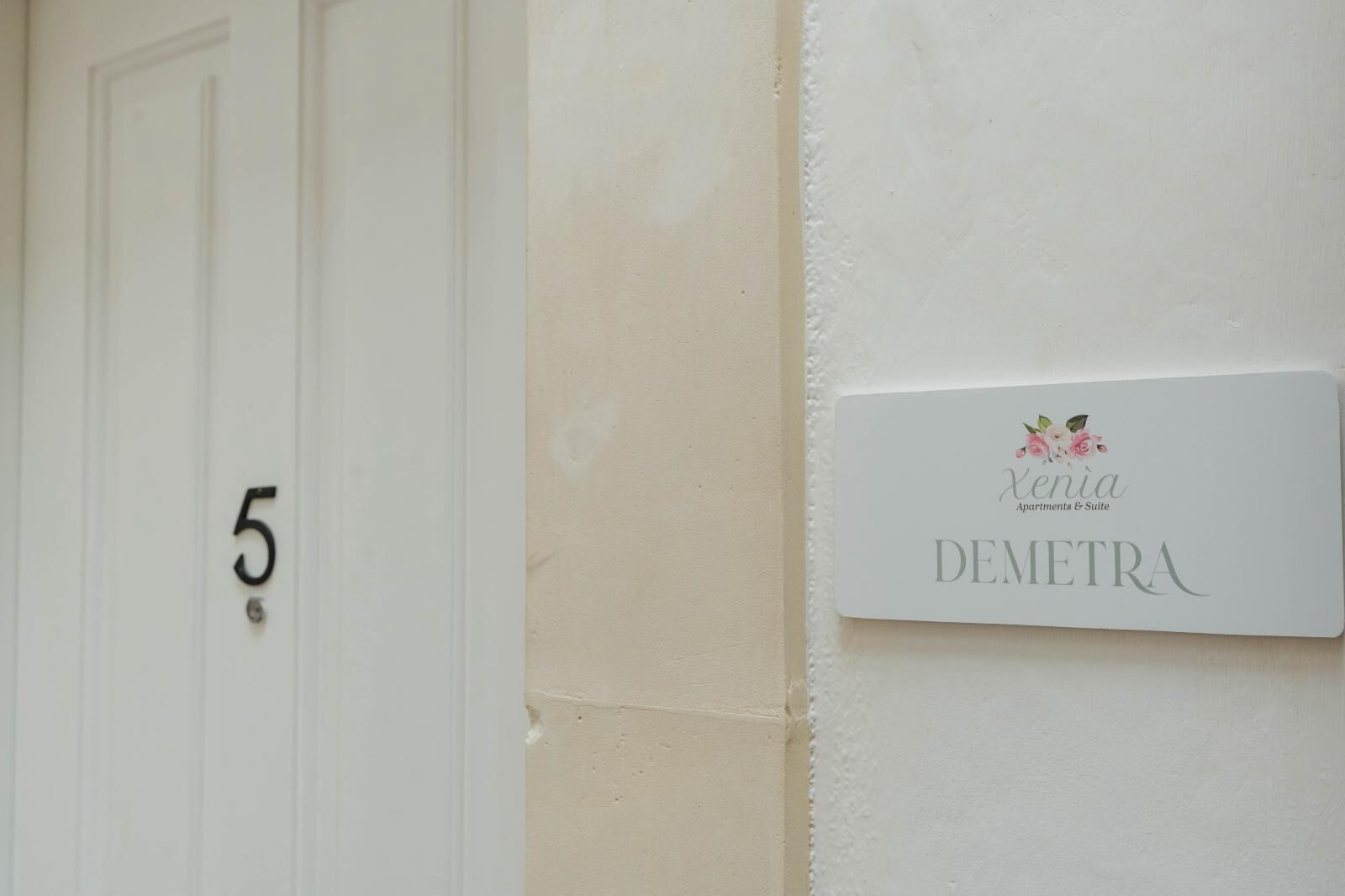 Une porte blanche portant le numéro 5 et une enseigne sur le mur indiquant : « Xenia Apartments & Suite DEMETRA ».