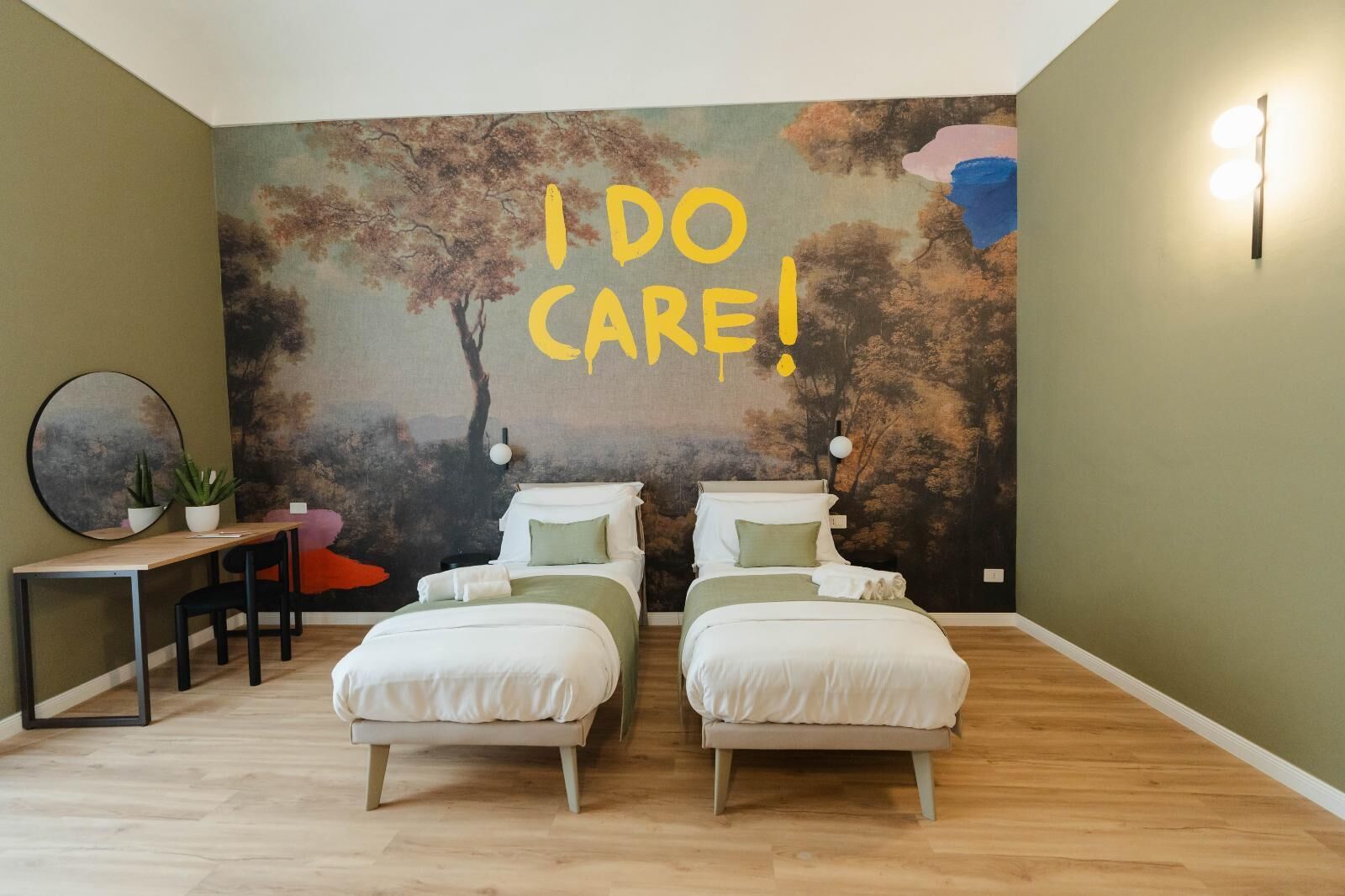 Une chambre avec deux lits, un bureau et une chaise. Un grand mur est décoré d’une fresque sur laquelle on peut lire le texte : « J’Y TIENS ! »
