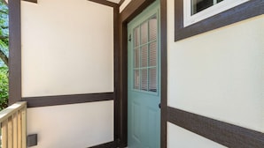 Una esquina de un edificio con pared blanca y detalles oscuros, una puerta verde con contraventanas y una ventana encima de la puerta.