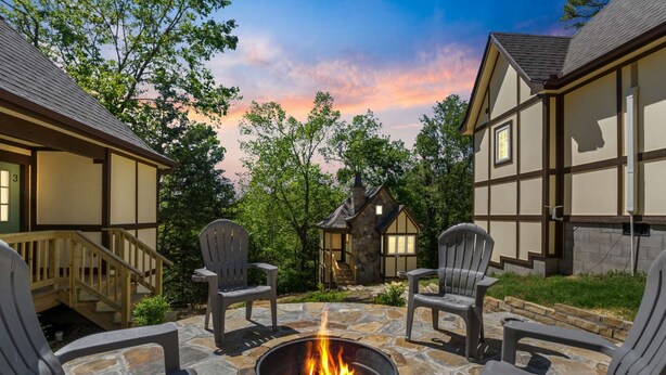 Un rincón al aire libre acogedor, con un hogar de piedra rodeado de sillas Adirondack, frente a casas tradicionales con entramado de madera y abundante vegetación.