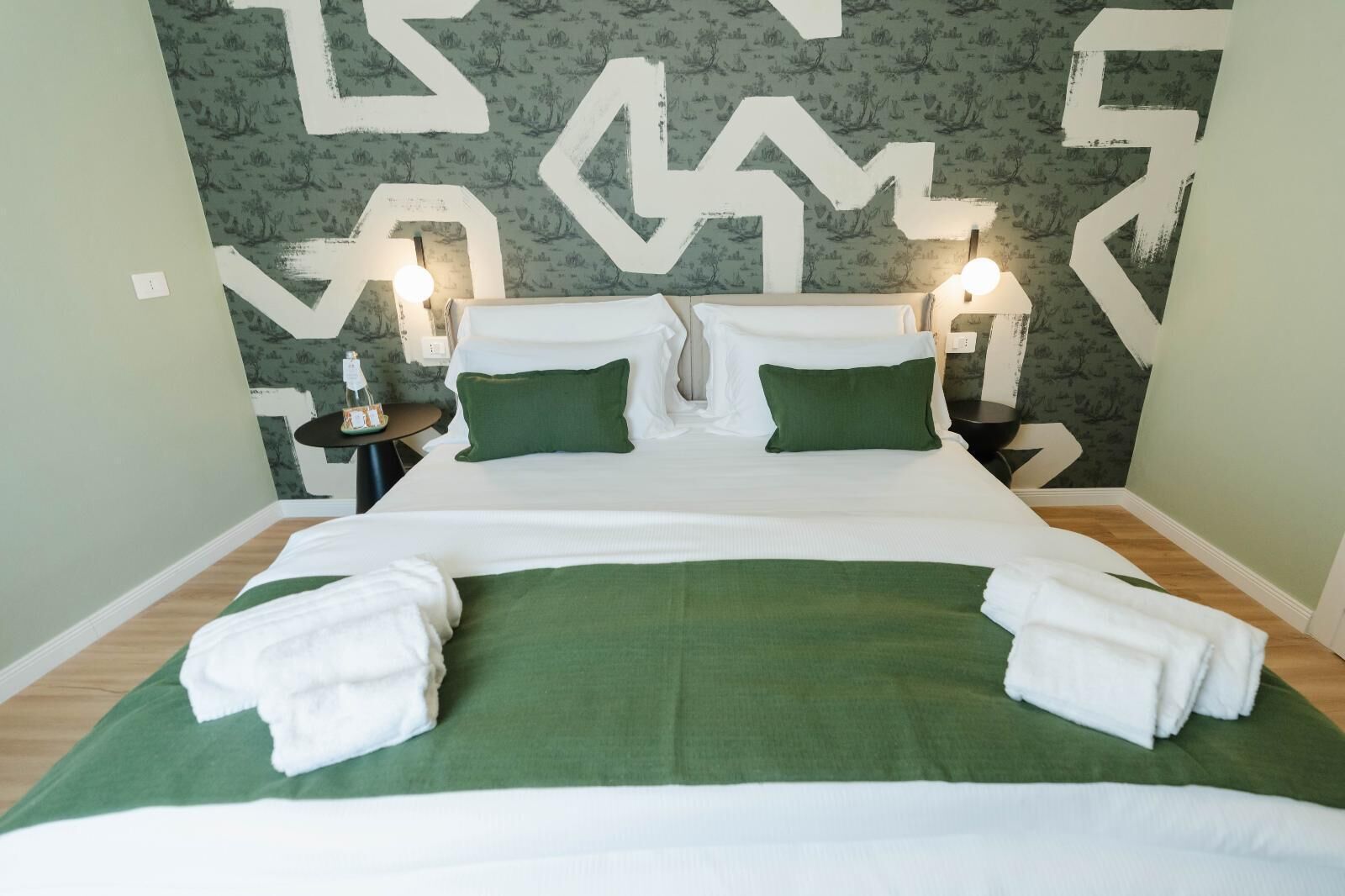 Ein Doppelbett mit grüner Bettwäsche und weißen Kissen, zwei Nachttische mit Lampen und eine Wand mit Muster im Hintergrund.