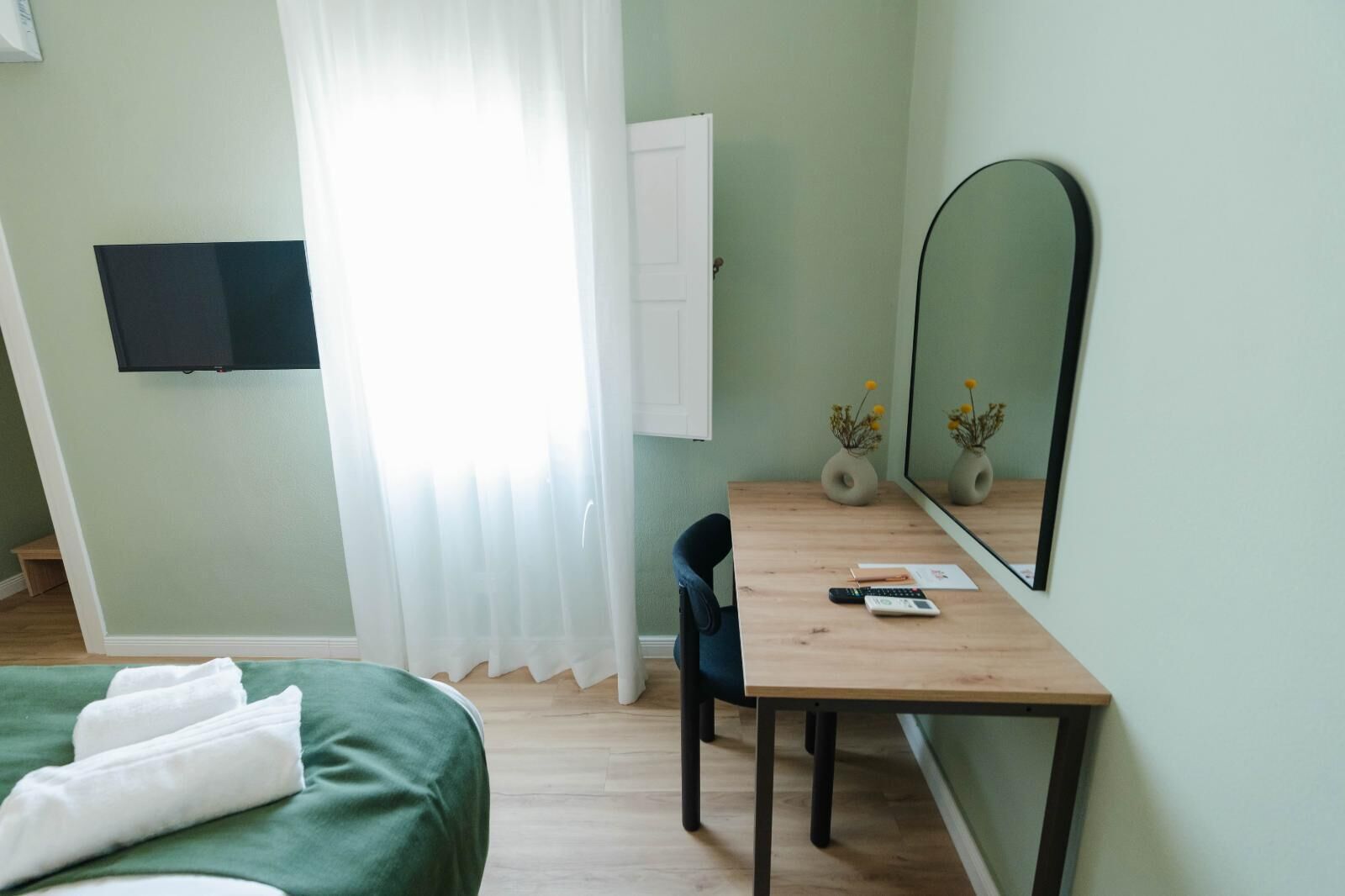 A hotel room with a bed, a desk with a chair, a flat-screen TV, a mirror, and two vases with flowers.