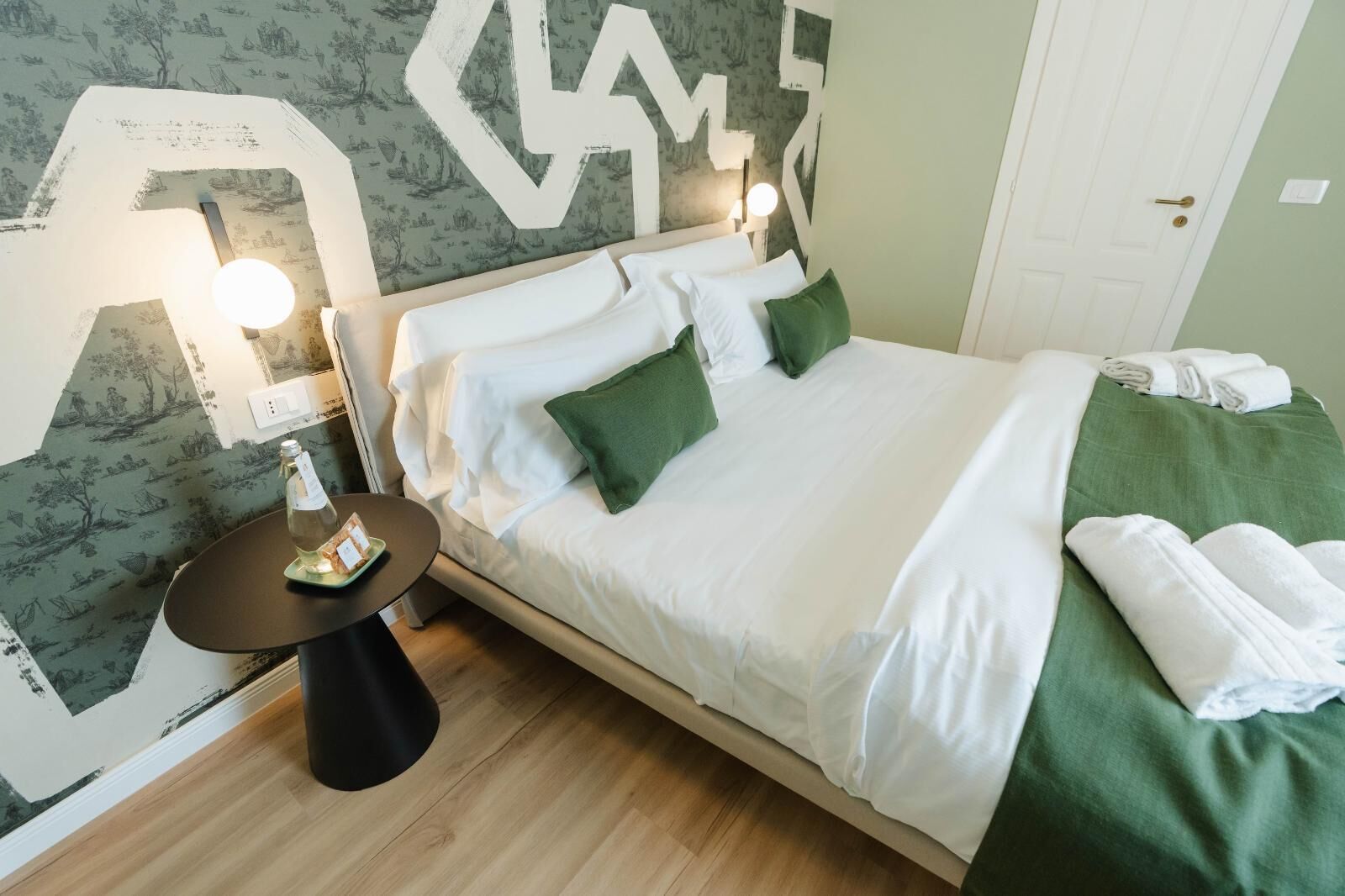 A neatly made bed with white and green bedding, a bedside table with a bottle and a plate, and a wall with a large white letter 'M'.