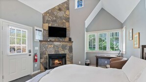A bedroom with a stone fireplace, a bed, a desk with a chair, and a window with blinds. - Hearthstone Hideaway at Tudor Suites (Eureka Springs)