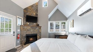 A bedroom with a stone fireplace, a bed with white bedding, a desk with a chair, and a window with blinds.