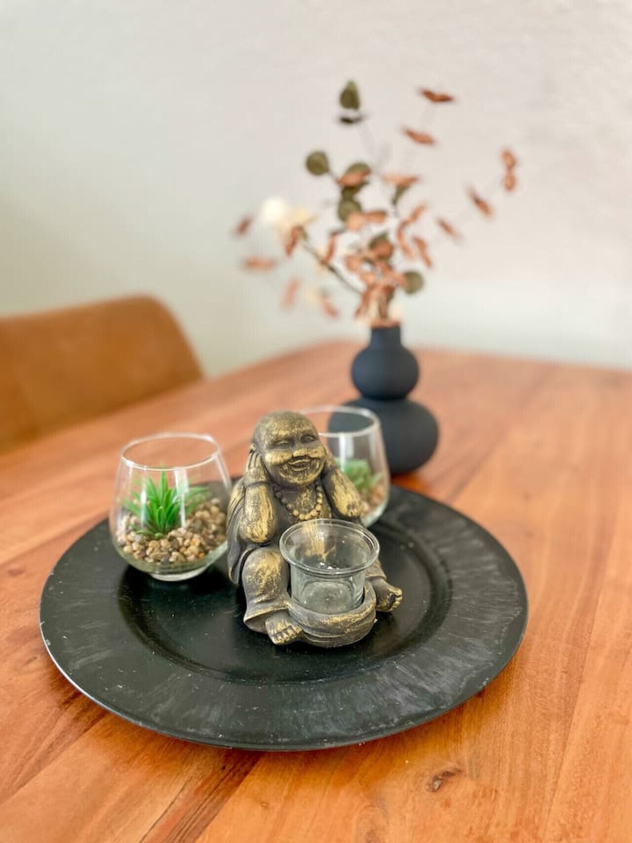 En sort tallerken med to små glasbeholdere og en Buddha-figur på et træbord.
