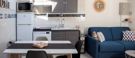 A compact kitchen with a microwave, refrigerator, and dishwasher. There's a dining table with chairs and a blue sofa with patterned cushions.