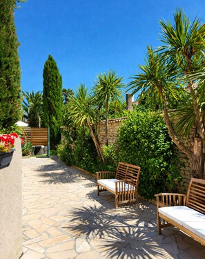 A patio with stone flooring, wooden benches, and lush greenery including palm trees and a tall conifer. - Le Clos des Sternes (La Flotte)