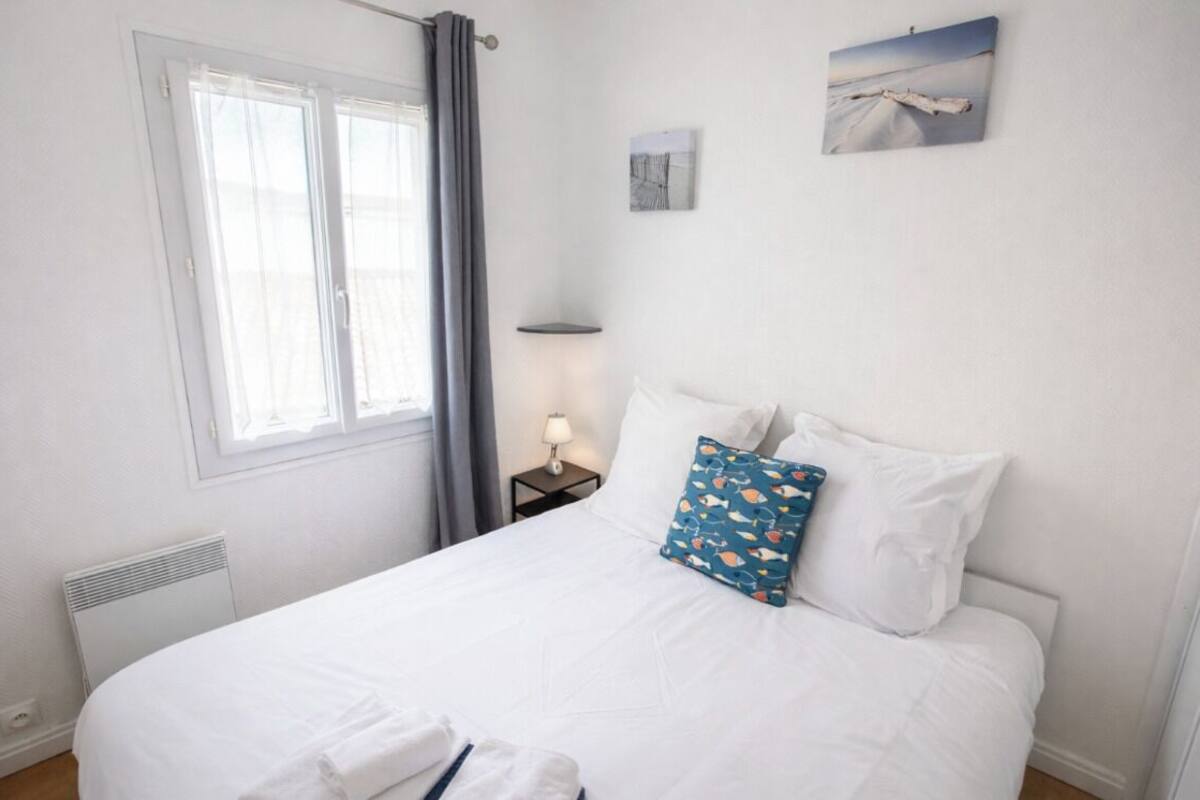 Ein ordentlich bezogenes Bett mit weißer Bettwäsche und einem einzelnen Zierkissen, ein Nachttisch mit Lampe, ein Fenster mit Vorhängen und zwei an der Wand hängende Bilder.