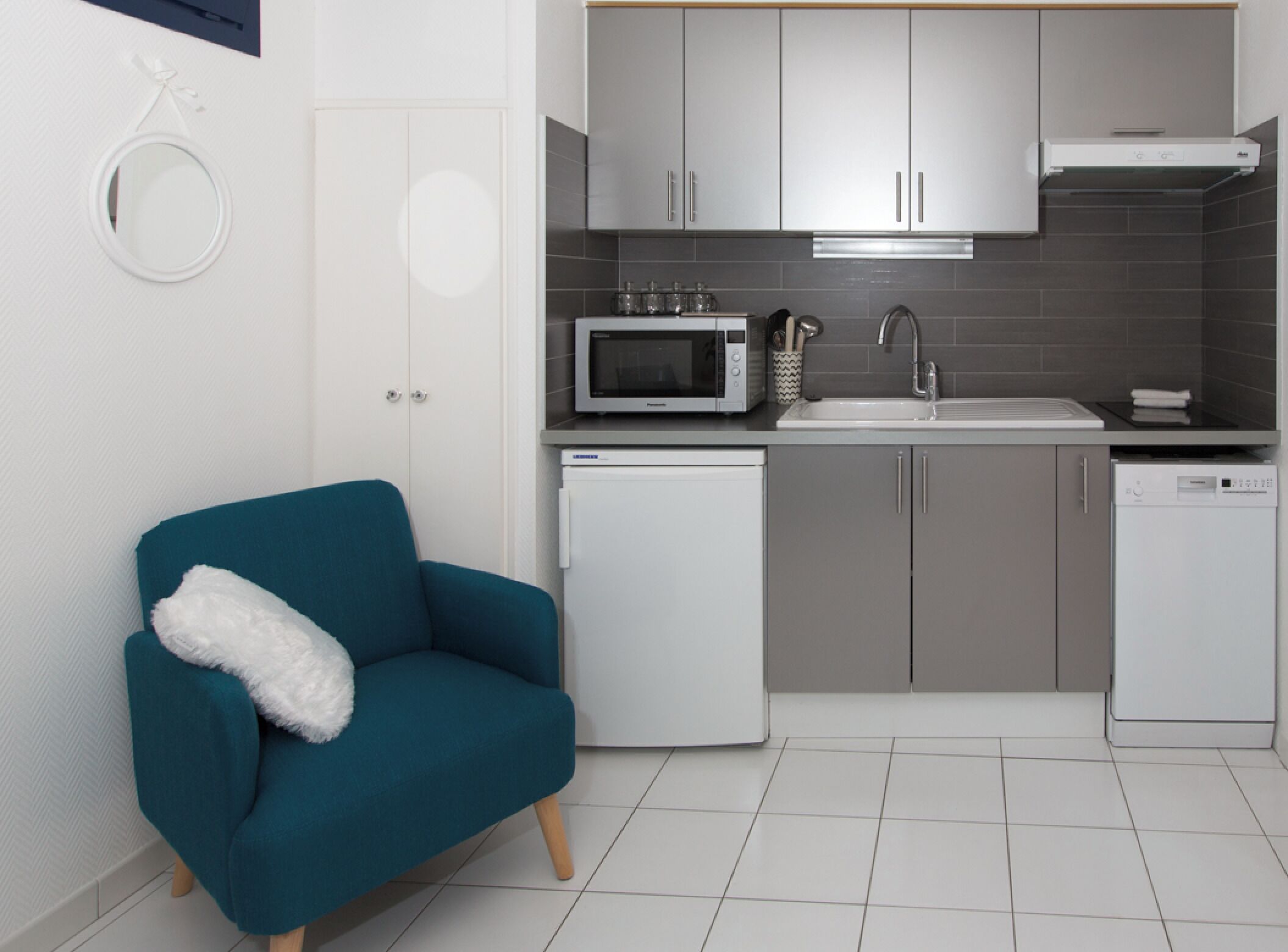 A compact kitchen with a microwave, sink, and dishwasher, a teal sofa with a white throw, and a small refrigerator.