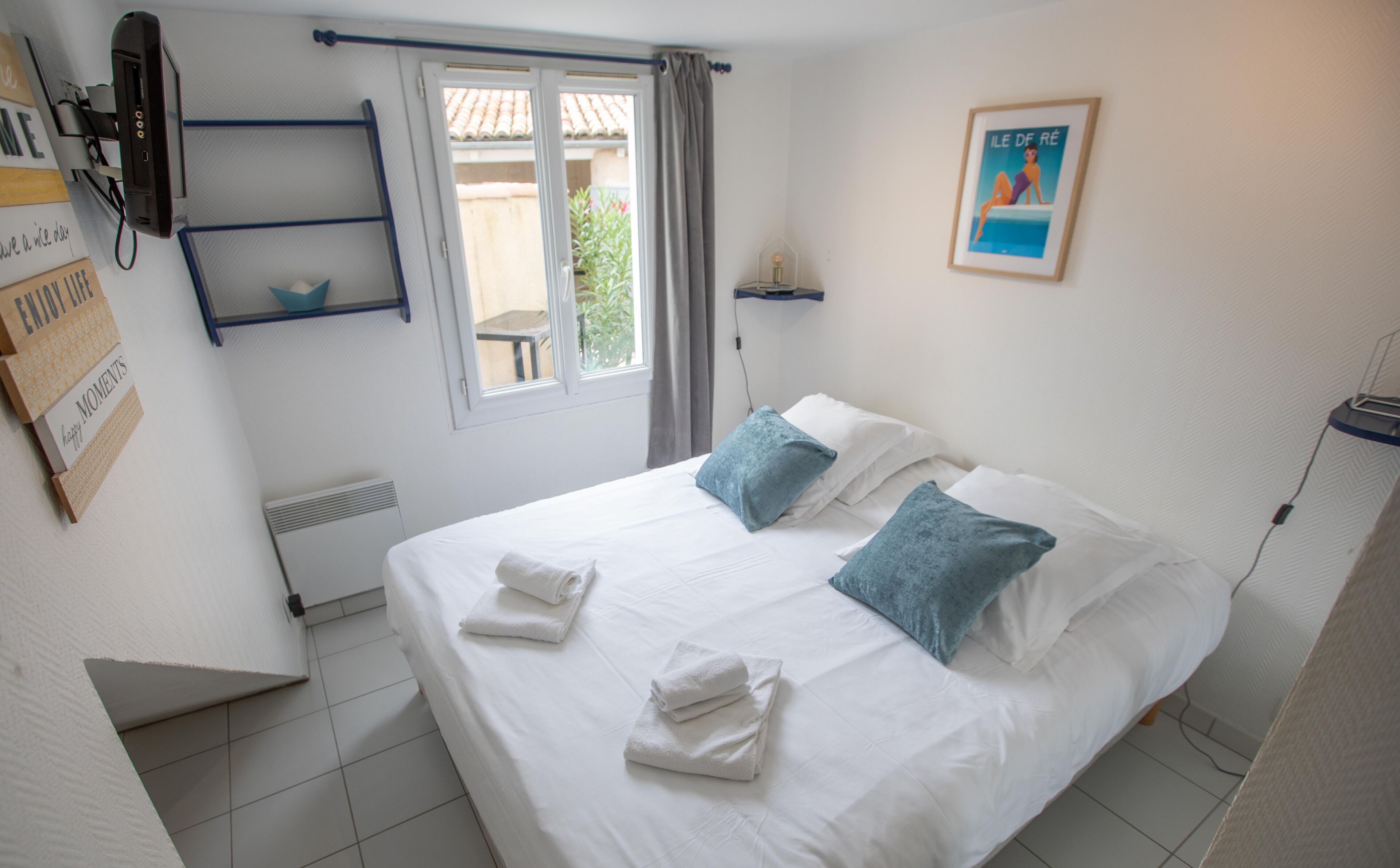 Una cama bien hecha con ropa blanca y dos almohadas azules, una mesita con una silla cerca de la ventana, y un cuadro en la pared.