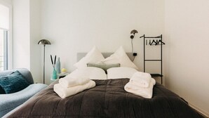 Ein ordentlich bezogenes Bett mit einer dunkelbraunen Tagesdecke und weißen Kissen, ein Nachttisch mit Lampe und Dekoration sowie ein an der Wand befestigter Kleiderhaken.