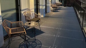 A covered outdoor seating area with wicker chairs and tables, adjacent to a building with large windows and a brick facade.